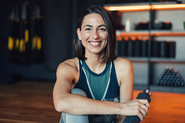 Mulher sorridente em um ambiente de academia, sentada e segurando uma garrafa de água, ideal para ilustrar vida saudável e atividades físicas.