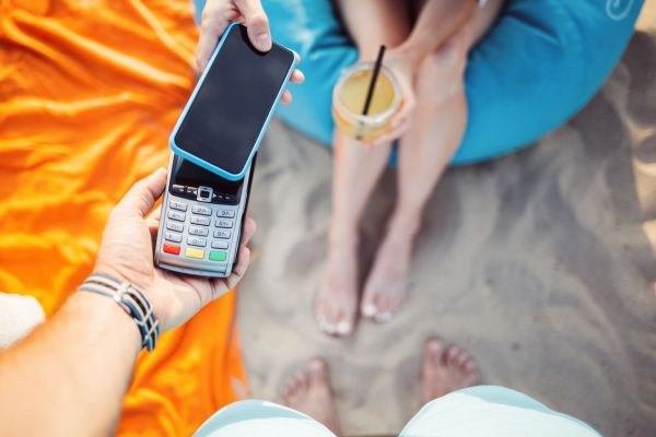 Pessoa na praia passando um cartão em uma maquininha, enquanto segura um copo com bebida.
