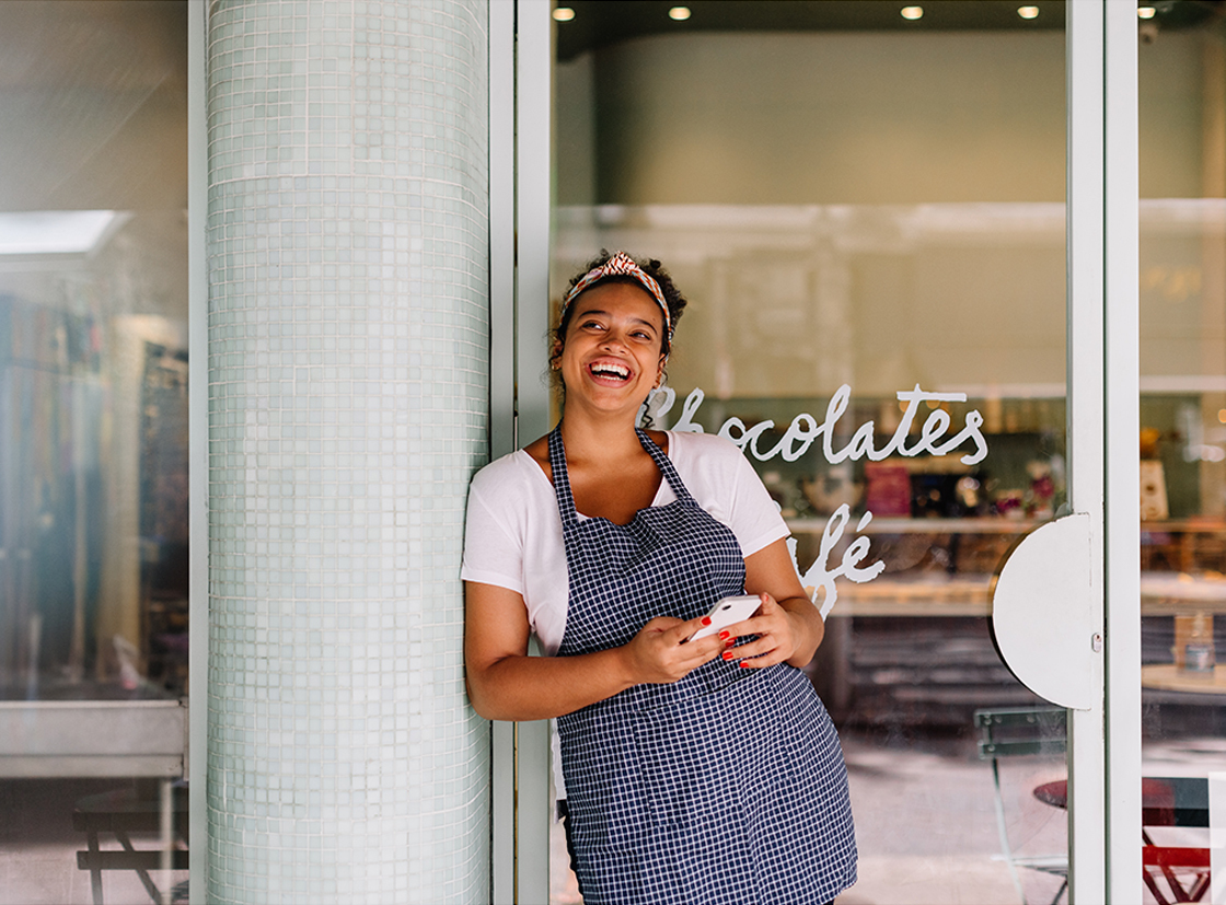 Mulher sorridente usando avental em frente a uma loja de chocolates, segurando um celular e emanando alegria. Perfeita para destacar a cultura de chocolates.