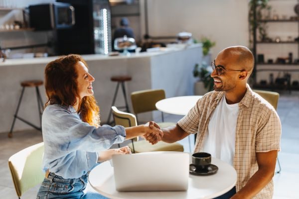 Dois empresários sentados à mesa, apertando as mãos em sinal de acordo, com expressões confiantes, simbolizando parceria e colaboração profissional.