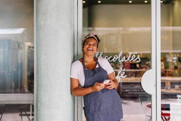 Empreendedora sorridente de avental azul e branco, encostada em uma porta de vidro de cafeteria, transmitindo felicidade e realização profissional