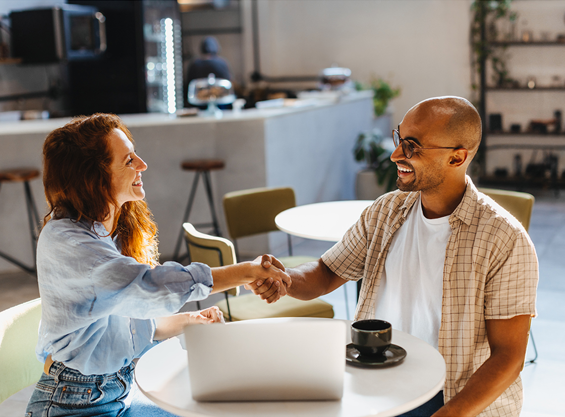 Dois profissionais felizes se cumprimentando em um ambiente de trabalho moderno, simbolizando parcerias e networking. A cena destaca um laptop e um café.