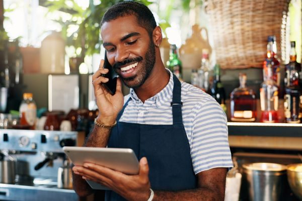 Homem empreendedor com avental azul, usando celular e tablet em um ambiente de café, representando eficiência e conectividade no gerenciamento do negócio.