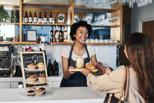 Imagem de uma barista sorridente servindo cappuccino e croissants a uma cliente em uma cafeteria. O ambiente é acolhedor e moderno, com bolos e bebidas ao fundo.