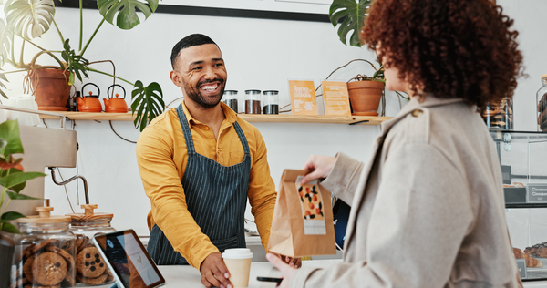 Atendente sorridente em cafeteria, entregando produtos para cliente em ambiente acolhedor. Destaca relação calorosa entre atendente e cliente.