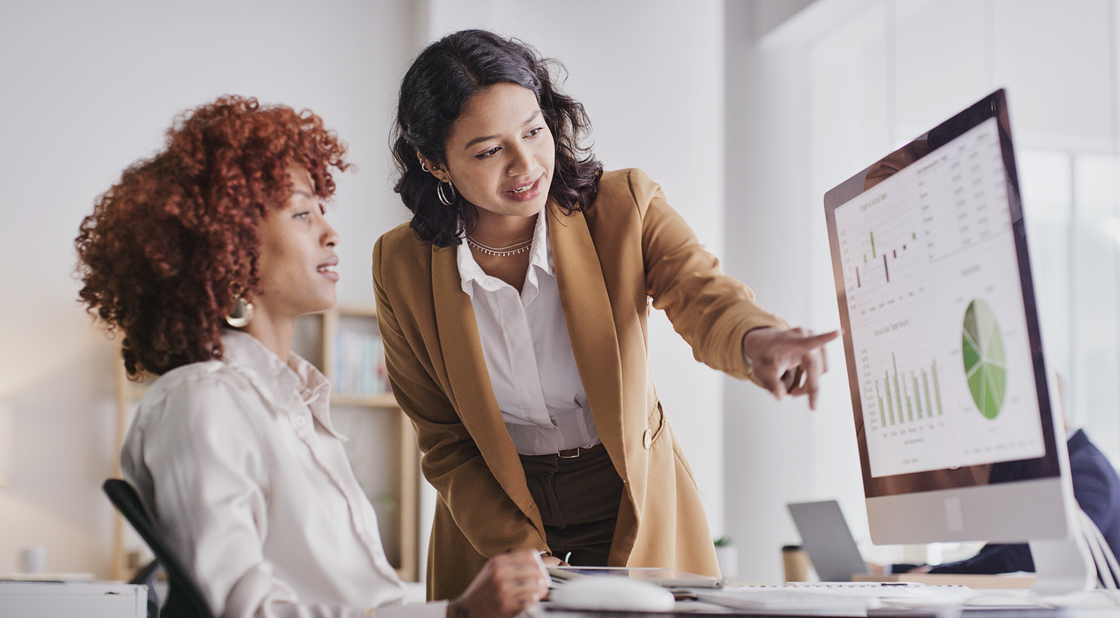 Duas mulheres em um ambiente de escritório analisando gráficos em um computador. Uma delas aponta para os dados na tela, demonstrando colaboração e trabalho em equipe.