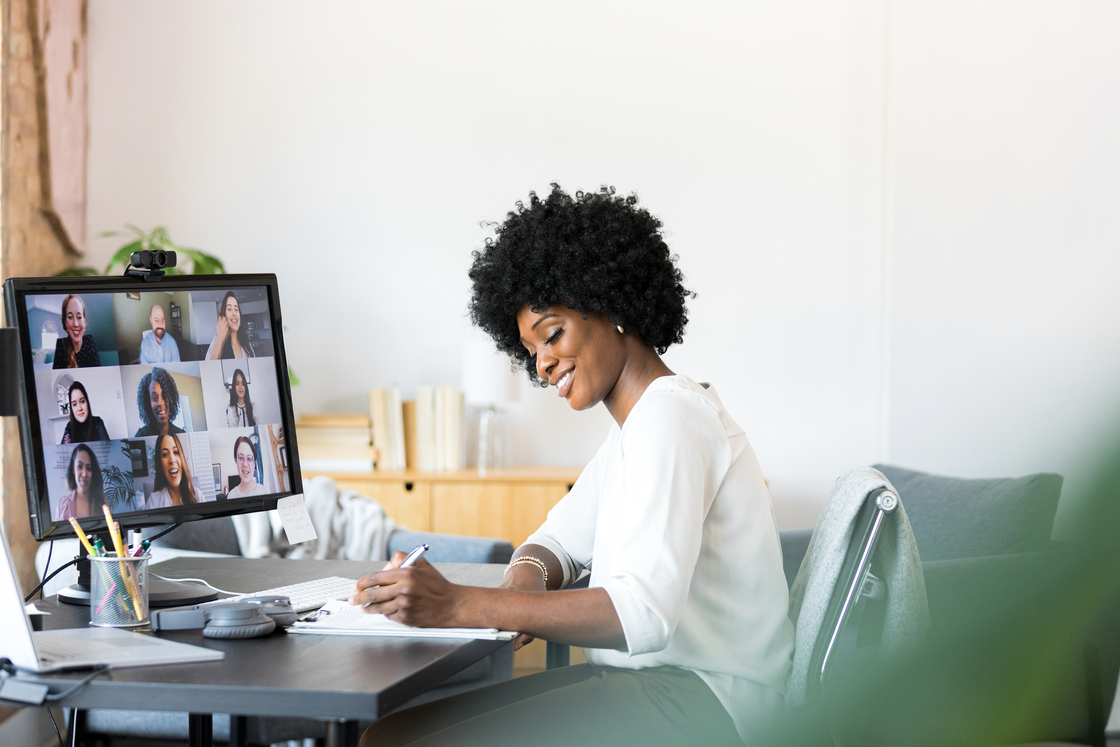 Mulher com cabelo cacheado sorrindo enquanto participa de uma videoconferência, anotando informações em um bloco de notas. Ambiente home office.