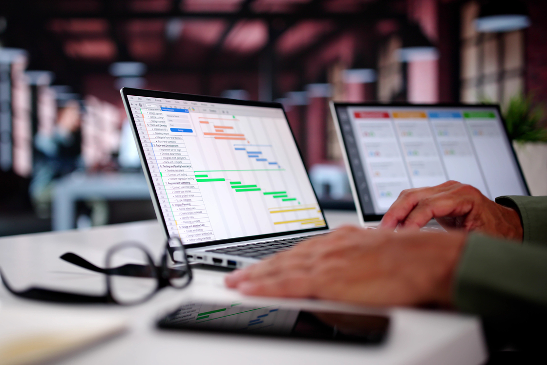 Pessoa utilizando um laptop com gráficos de planejamento em um ambiente de trabalho. A tela mostra um cronograma com várias cores, destacando a organização de tarefas e projetos.
