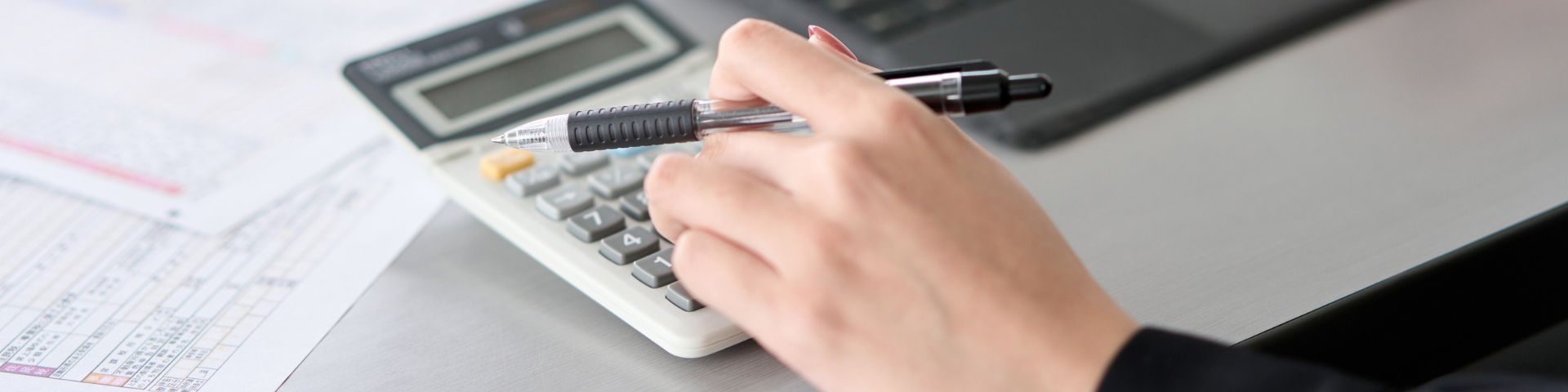 Mão com caneta sobre calculadora e planilhas, calculando tributos na nota fiscal fora do Simples Nacional.