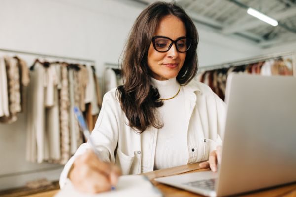 Mulher trabalhando em um escritório de moda, escrevendo à mão enquanto usa um laptop, cercada por roupas em um cabideiro.