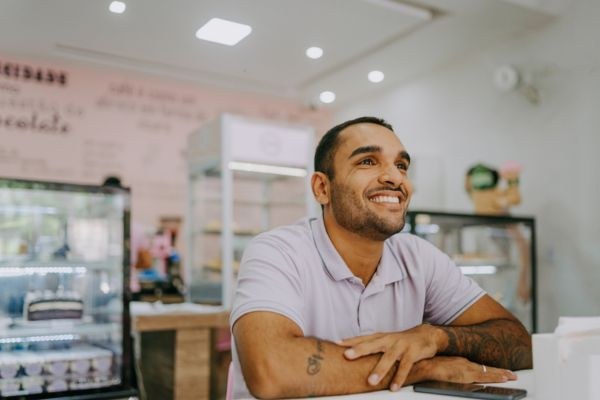 Homem sorridente em uma confeitaria, transmitindo alegria e satisfação. Ele está vestindo uma camisa azul e parece animado em um ambiente acolhedor.
