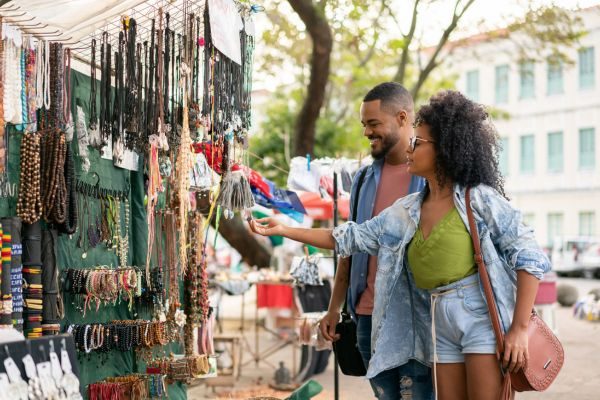 Casal sorridente visitando uma feira ao ar livre, onde a mulher observa acessórios artesanais expostos em uma barraca, com árvores e prédios ao fundo.
