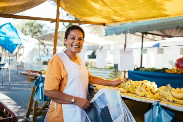 Feirante sorridente em sua barraca, organizando bananas e outros produtos frescos, com uma lona amarela ao fundo protegendo do sol.